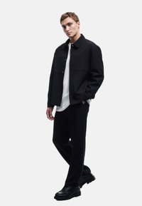 Veste noire à boutons avec deux poches avant, portée sur une chemise blanche. Associée à un pantalon noir et des chaussures noires épaisses. Texture de tissu lisse.