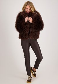 Bruine, harige nep-vachtjas met een boxy pasvorm, gecombineerd met zwarte leren broek en bruine sneakers met witte strepen.