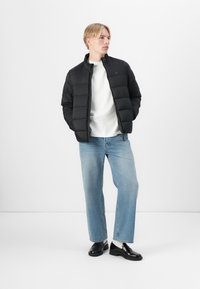 Jachetă puffer neagră cu textură matlasată, guler înalt, asociată cu o cămașă albă și blugi ușor albaștri. Pantofi negri lustruiți completează ținuta.