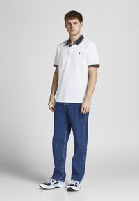 Jauns vīrietis stāv, ģērbies baltā polo krekliņā ar tumši zilām apkaklītēm, zilās džinsās un baltos sporta apavos uz vienkrāsaina pelēka fona.