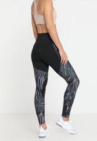 Una donna che indossa leggings neri e grigi a fantasia e un reggiseno sportivo beige, in piedi con una gamba leggermente sollevata su uno sfondo bianco.