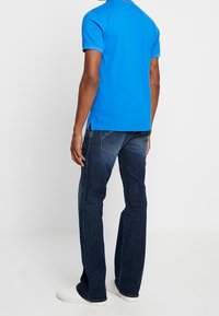 Camisa polo azul royal de manga corta, vista trasera, combinada con jeans de mezclilla azul oscuro y zapatillas blancas. Diseño simple con aberturas laterales.