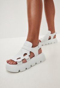 White platform sandals with cut-out design, thick sole, and adjustable strap. Made of smooth material, featuring a rounded toe and open sides.