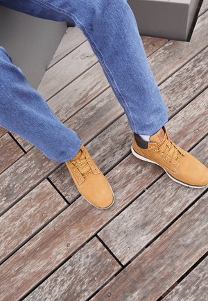 Person wearing blue jeans and tan lace-up boots sitting on a gray bench on a wooden deck floor.