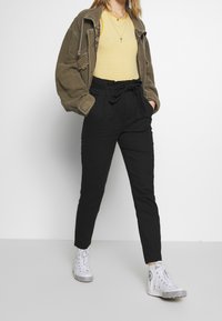 Černé kalhoty s vysokým pasem, zavázané v pase, kombinované se žlutým pruhovaným topem a olivově zelenou oversized bundou. Bílé tenisky s vzorem na podrážce.