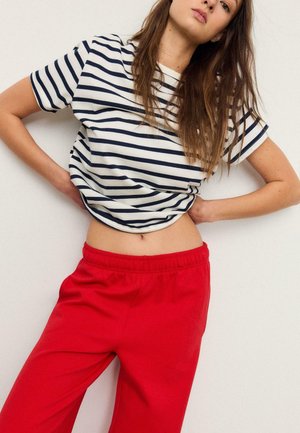 Frau trägt ein marineblau-weiß gestreiftes Shirt, hochgezogen, um den Bauch freizulegen, kombiniert mit leuchtend roten Hosen, steht vor einem einfarbigen Hintergrund.