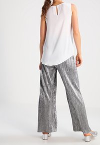 Blusa bianca senza maniche con dettaglio a chiave sulla schiena, abbinata a pantaloni a vita alta plissettati argentati lucidi e scarpe metalliche open-toe.