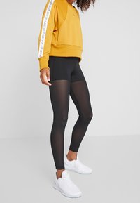 Czarne legginsy z przezroczystymi panelami, zestawione z musztardowym sweatshirt'em z białym napisem "Just Do It" na rękawach, noszone z białymi trampkami.