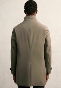 Khakifarbener Trenchcoat mit hohem Kragen, einer einzigen Naht auf der Rückseite und geknöpften Manschetten, aus einem glatten, leichten Stoff gefertigt.
