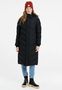 Lang sort dunfrakke med chevronmønster, lynlås-lommer og hætte. Båret med lyseblå jeans og sorte klodsede ankelstøvler. Rød hue på hovedet.