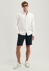 Camisa de lino blanca, pantalones cortos de textura navy cortos, zapatillas casuales con acentos en gris y blanco, y gafas de sol negras redondas.