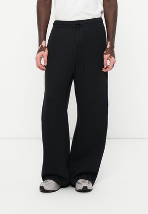 Personne portant un pantalon noir large à cordon, une chemise blanche et des baskets grises, debout devant un fond blanc uni.