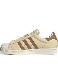 Beige leather sneaker with three brown stripes, rubber toe cap, and textured brown heel accent. Features a padded collar and lacing system.