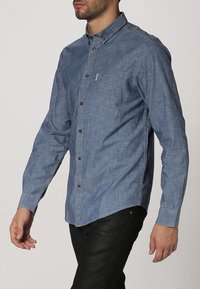 Camisa azul claro de manga larga con botones en tela chambray. Cuenta con un bolsillo en el pecho y botones marrones en el frente, textura suave.
