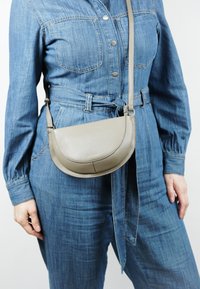 Grijze leren schoudertas met een gebogen vorm, een gladde textuur en een verstelbare schouderriem, gedragen met een denim jumpsuit.