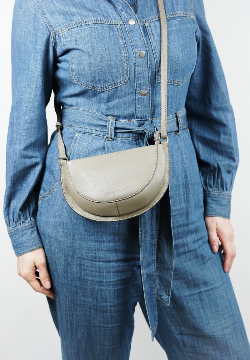 Grijze leren schoudertas met een gebogen vorm, een gladde textuur en een verstelbare schouderriem, gedragen met een denim jumpsuit.