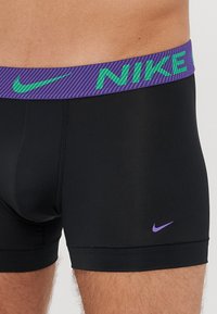 Črne športne hlačke z elastičnim pasom v vijoličnih in zelenih črtah, ki prikazuje logo "NIKE", gladka tekstura in prilegajoč dizajn.