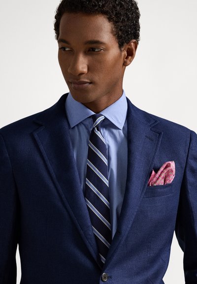 Vyriškis dėvi mėlyną švarko švarką, šviesiai mėlyną marškinius, dryžuotą tamsiai mėlyną kaklaraištį ir rožinį kišeninį skudurėlį su raštais, žiūrintis į šoną balto fono atžvilgiu.