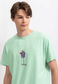 T-shirt en coton vert clair avec un col rond, présentant un graphique coloré d'une personne prenant une photo au centre. Texture douce.