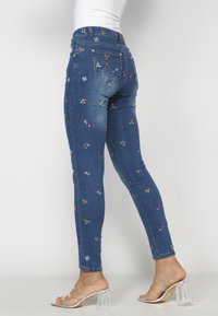 Slim-fit džíny v tmavě modré barvě s barevným květinovým vyšíváním. Mají střední pas a standardní design se pěti kapsami.