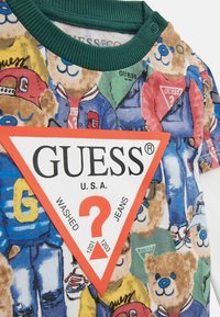 Marškinėliai su žaliu banguotu apykakle, puoštas spalvingu lokio raštu ir dideliu raudonu trikampiu logotipu su užrašu "GUESS U.S.A." ir klausimo ženklu.