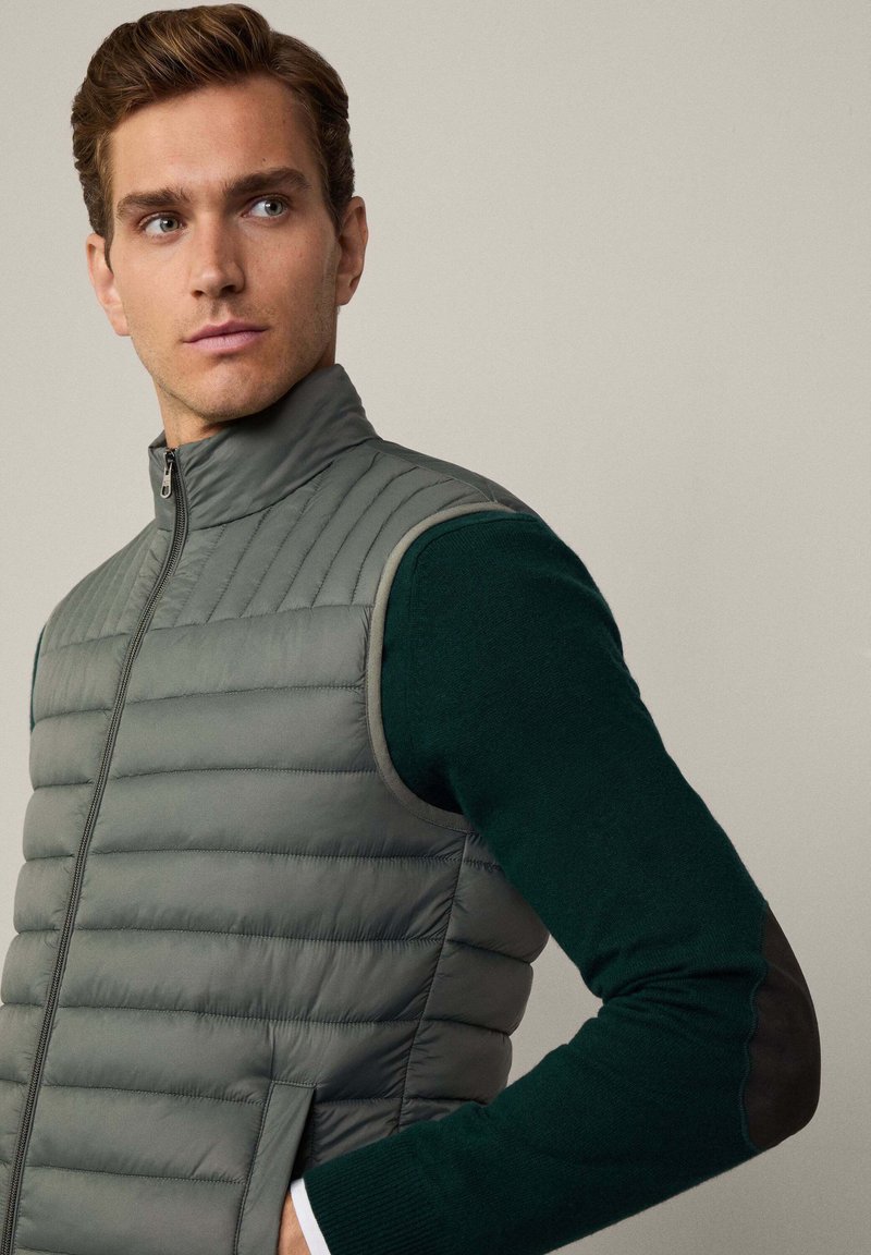 Hellgraue gepolsterte Weste mit hohem Kragen und Reißverschluss, getragen über einem dunkelgrünen Pullover mit schwarzen Ellbogen-Patches.