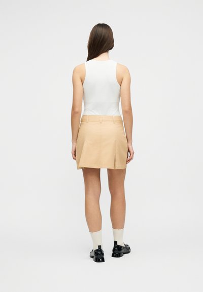 Femme portant un haut blanc sans manches, une jupe plissée beige, des chaussettes blanches et des chaussures noires, debout de dos sur un fond uni.