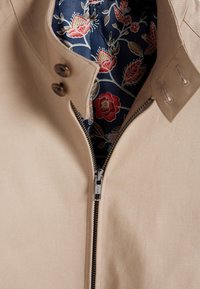 Beige jakke med krave, med en metal lynlås. Indersiden viser marineblåt stof med blomster mønstre i rød, pink og grøn.