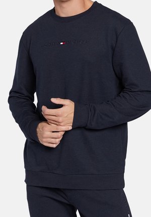 Mann trägt dunkelblaue Tommy Hilfiger Sweatshirt mit Logo auf der Brust, die Hände vor der Taille verschränkt.