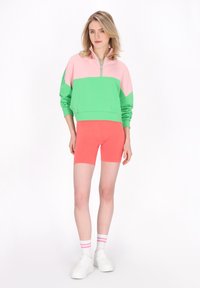 Sudadera de cremallera con bloques de color en verde y rosa, combinada con pantalones cortos de ciclismo coral ajustados. Calcetines blancos con rayas rosas, zapatillas blancas.