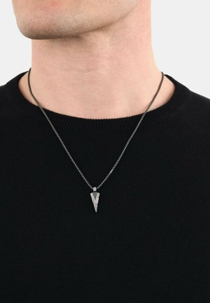 Collana con pendente in metallo argentato caratterizzata da un design triangolare, appesa a una catena sottile, indossata sopra un maglione nero con texture.