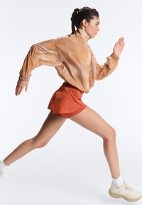 Femme athlétique aux cheveux tressés courant, portant un coupe-vent orange, un short couleur rouille, des chaussettes blanches et des chaussures de course beiges.