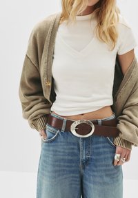 Svetlohnedý kardigán cez biely top s krátkym rukávom, kombinovaný so svetlomodrými džínami. Tmavohnedý opasok so striebornou oválnou sponou a prsteňmi.