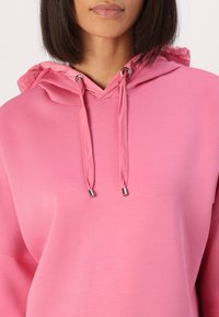 Felpa rosa con cappuccio caratterizzata da un cordino con estremità in argento, vestibilità rilassata e una trama morbida. Il cappuccio presenta un tessuto interno a contrasto.