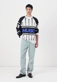 Overdimensjonert svart-hvit strikkegenser med blå "HUGO"-logo og geometrisk mønster, kombinert med lyseblå rette jeans og svarte sko.