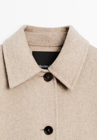 Beige wool coat with a pointed collar, featuring a button closure and a textured finish. Black label visible inside collar.