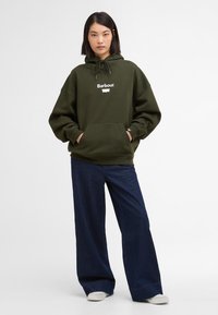 Ελιές πράσινη φούτερ με κουκούλα και μπροστινή τσέπη, που εμφανίζει το λευκό λογότυπο "Barbour" στο στήθος. Συνδυάζεται με ναυτικές παντελόνες φαρδιές.