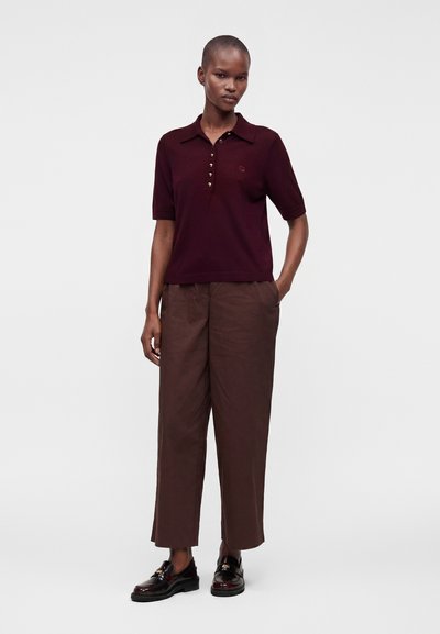 Personne debout, une main dans la poche, portant un polo à manches courtes bordeaux foncé et un pantalon marron ample avec des mocassins noirs.