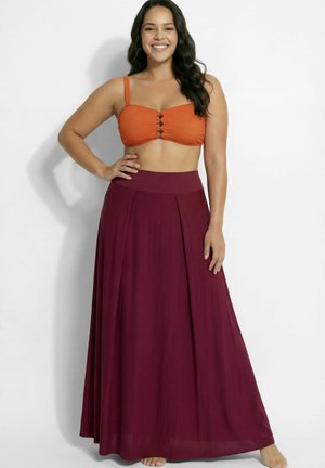 Lachende vrouw met hand op heup, draagt een oranje crop top met uitsparingen en een vloeiende bordeauxrode maxi-rok tegen een witte achtergrond.