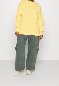 Gul crewneck sweatshirt med en liten logotyp, tillsammans med lösa, urtvättade gröna cargo-byxor och krämfärgade boots med tjock sula.