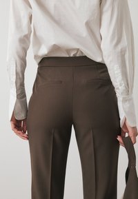 Bruine op maat gemaakte broeken met een gladde textuur, voorzien van twee achterzakken en een hoge tailleband, gecombineerd met een witte overhemd met knopen.
