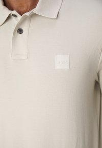 Beige polo shirt gemaakt van gestructureerd materiaal met een klassieke kraag, twee donkere knopen en een tonale vierkante logo op de linkerborst.