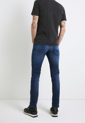 Homme aux bras tatoués portant un t-shirt noir, un jean bleu et des baskets noires, debout face à un mur blanc.
