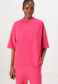 Rosa oversized t-skjorte med rund hals og korte ermer, med en subtil logo i en lysere tone, komplementert av matchende rosa bukser.