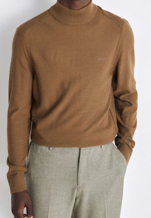 Homme portant un pull col montant marron avec la broderie "BOSS" sur la poitrine et un pantalon texturé gris clair, une main dans la poche.
