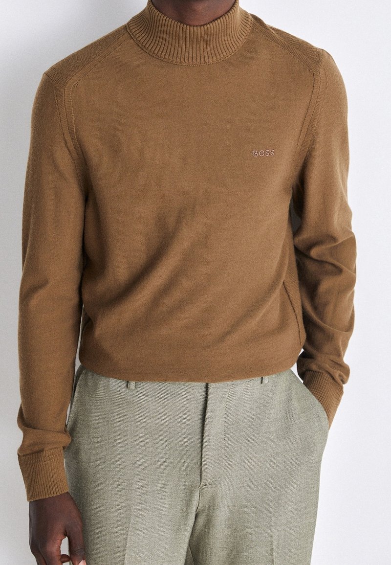 Homme portant un pull col montant marron avec la broderie "BOSS" sur la poitrine et un pantalon texturé gris clair, une main dans la poche.