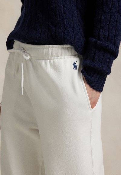 Person iført cremefarvede bukser med snører og et lille marineblåt polo-logo samt en marineblå ribstrikket sweater, hånden afslappet i lommen.