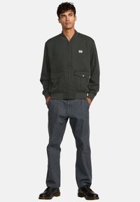 Giacca bomber verde scuro con due tasche frontali, polsini a coste e chiusura con zip. Indossata con pantaloni grigi e scarpe nere.