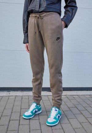 Person iført brune Nike joggingbukser, turkise og hvide Nike sneakers og en sort læderjakke, der står på gråt fortov.