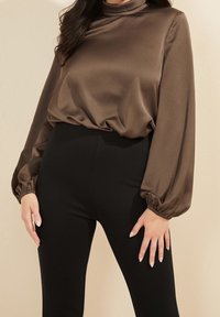 Brun satin langærmet top med høj hals, samlede manchetter og en afslappet pasform, parret med højtaljede sorte leggings.